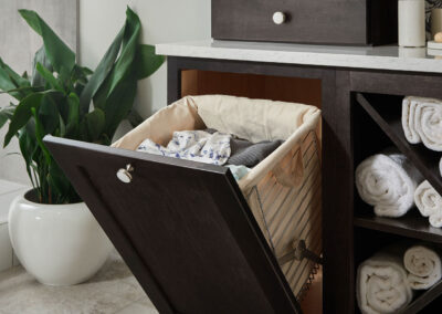 Tilt-out laundry hamper cabinet with a fabric-lined basket holding clothes, set in a dark wood vanity with rolled towels and a potted plant nearby.