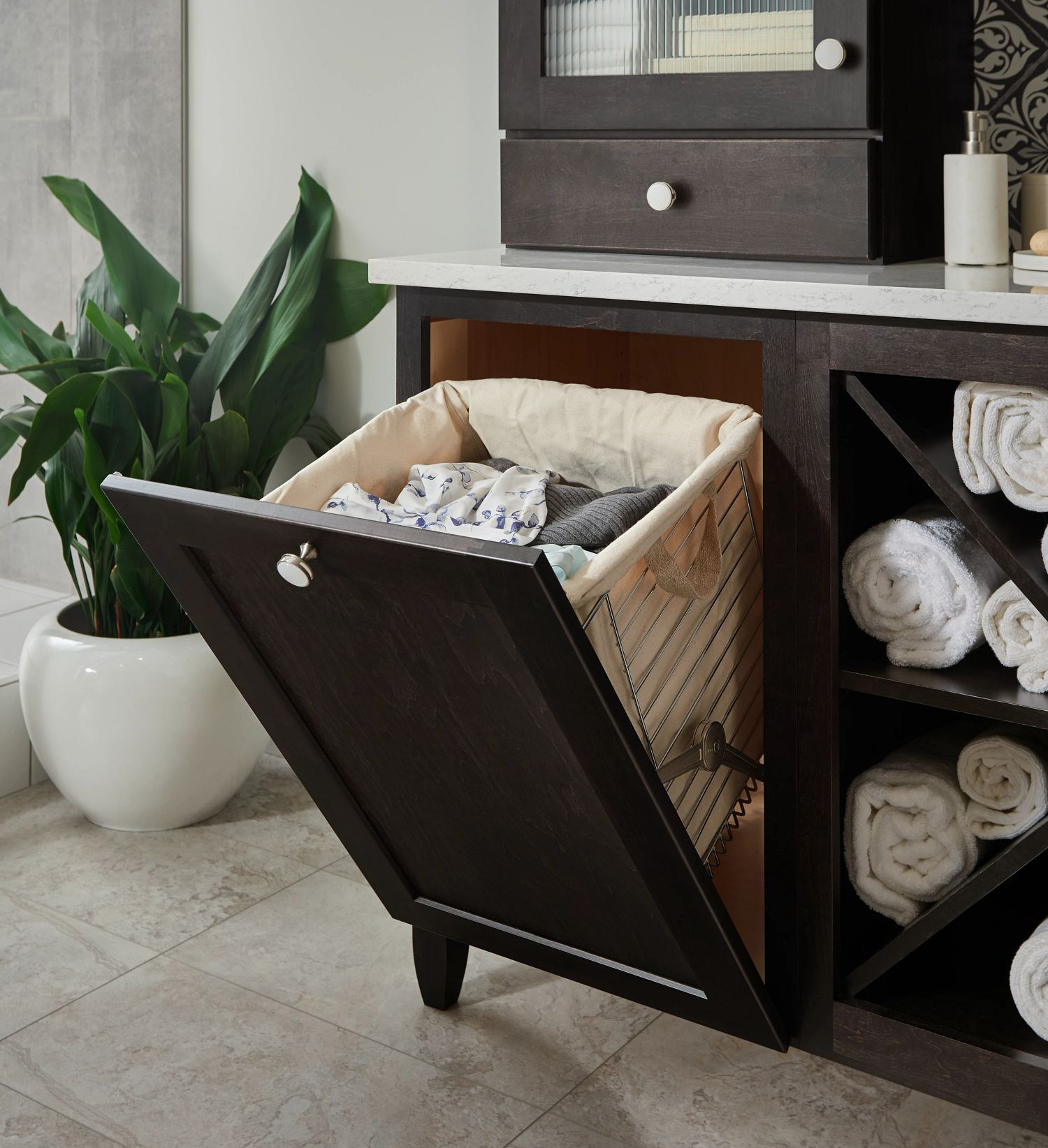 Tilt-out laundry hamper cabinet with a fabric-lined basket holding clothes, set in a dark wood vanity with rolled towels and a potted plant nearby.