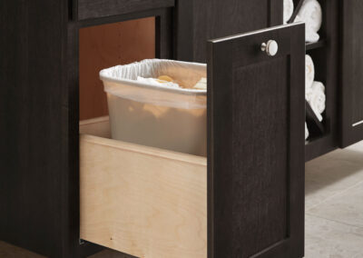 Pull-out trash bin cabinet with a plastic liner in a dark wood bathroom vanity with tiled flooring and patterned backsplash.