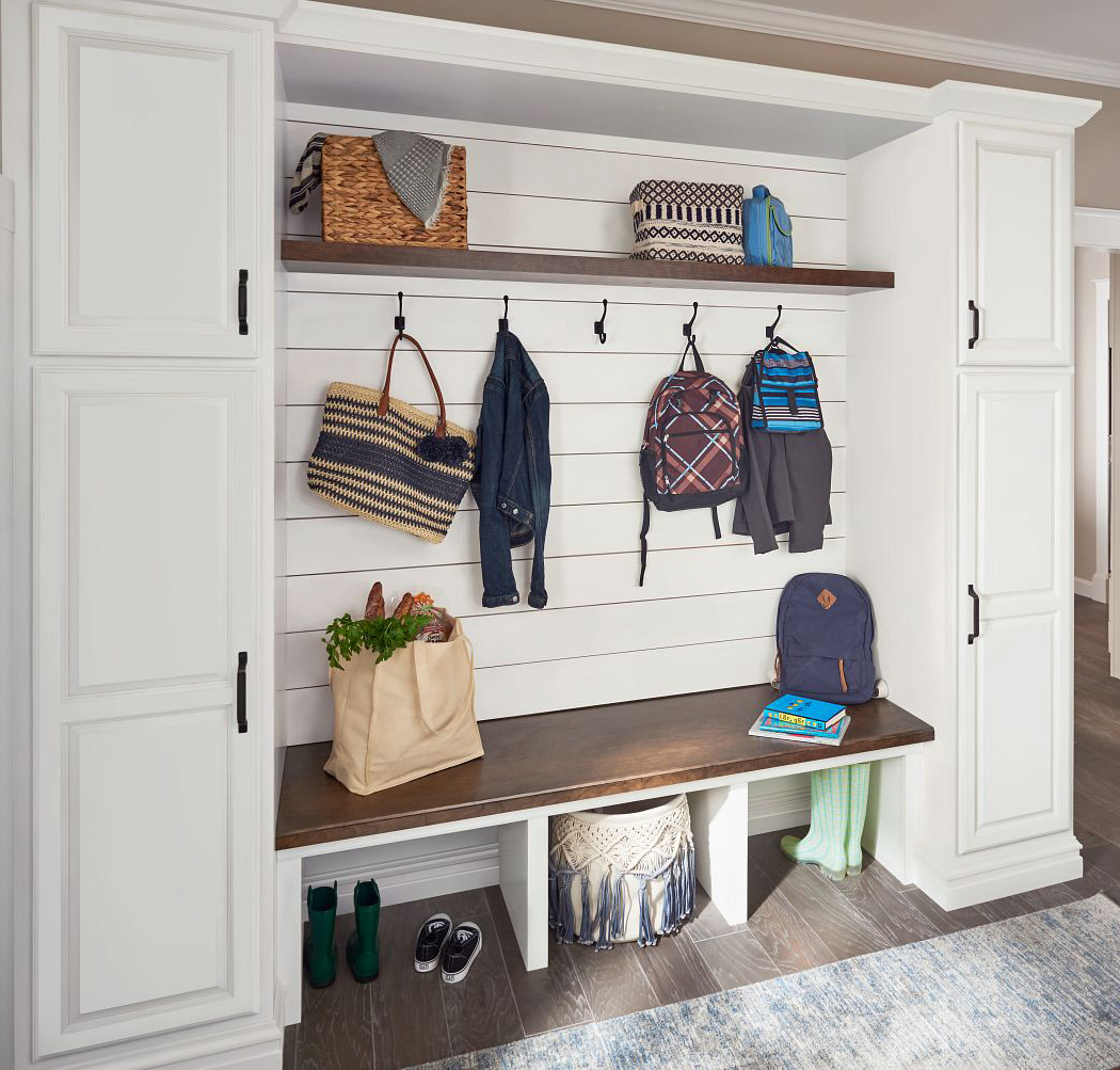 White mudroom storage unit with built-in bench, wall hooks holding bags and jackets, overhead shelf with baskets, and cubbies below for shoes and storage.