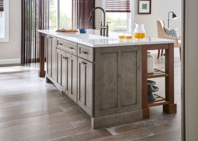 Kitchen island with gray wood-grain cabinets, white countertop, built-in sink and brushed metal faucet in a bright open kitchen with hardwood floors.