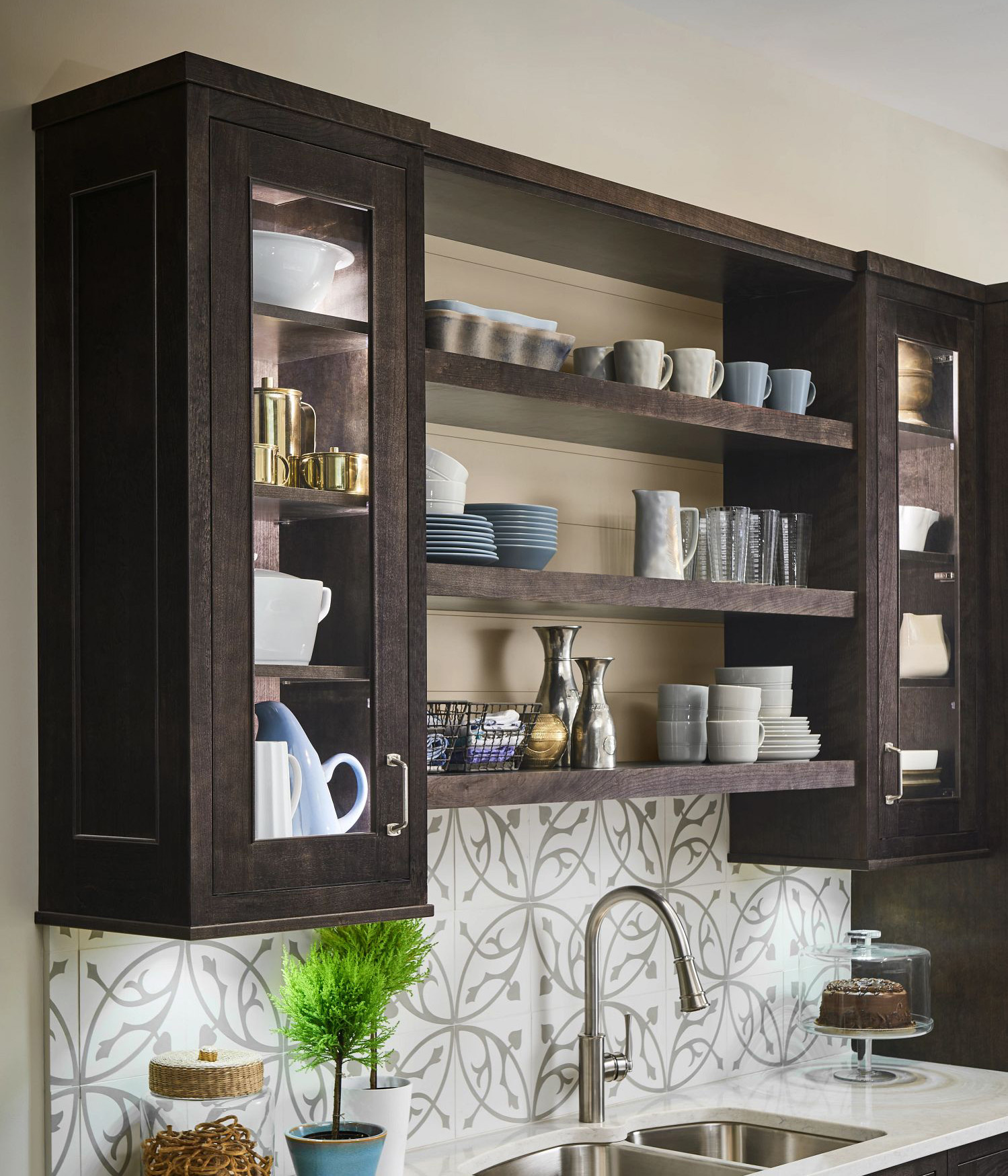 Modern kitchen with dark wood cabinets, open shelving displaying dishes and glassware, white countertop with double sink, patterned backsplash, and decorative items including potted plants and a cake stand.