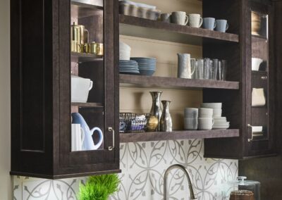 Modern kitchen with dark wood cabinets, open shelving displaying dishes and glassware, white countertop with double sink, patterned backsplash, and decorative items including potted plants and a cake stand.