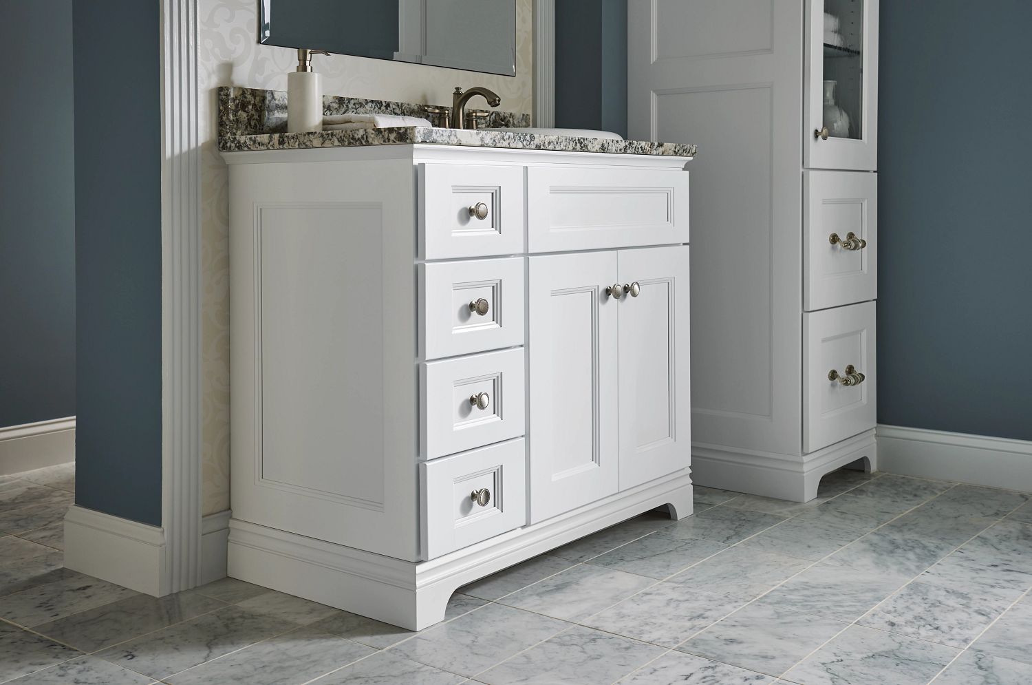 White bathroom vanity with granite countertop, silver hardware, and matching tall cabinet against blue walls and marble tile flooring.