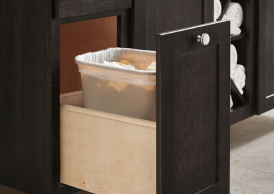 Pull-out trash bin cabinet with a plastic liner in a dark wood bathroom vanity with tiled flooring and patterned backsplash.
